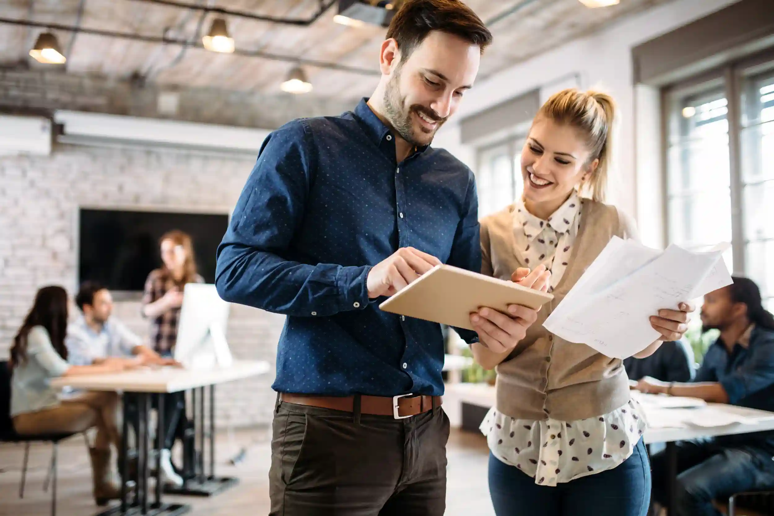 A man and woman stand in an office, one holding a tablet and the other holding papers, engaged in a discussion.