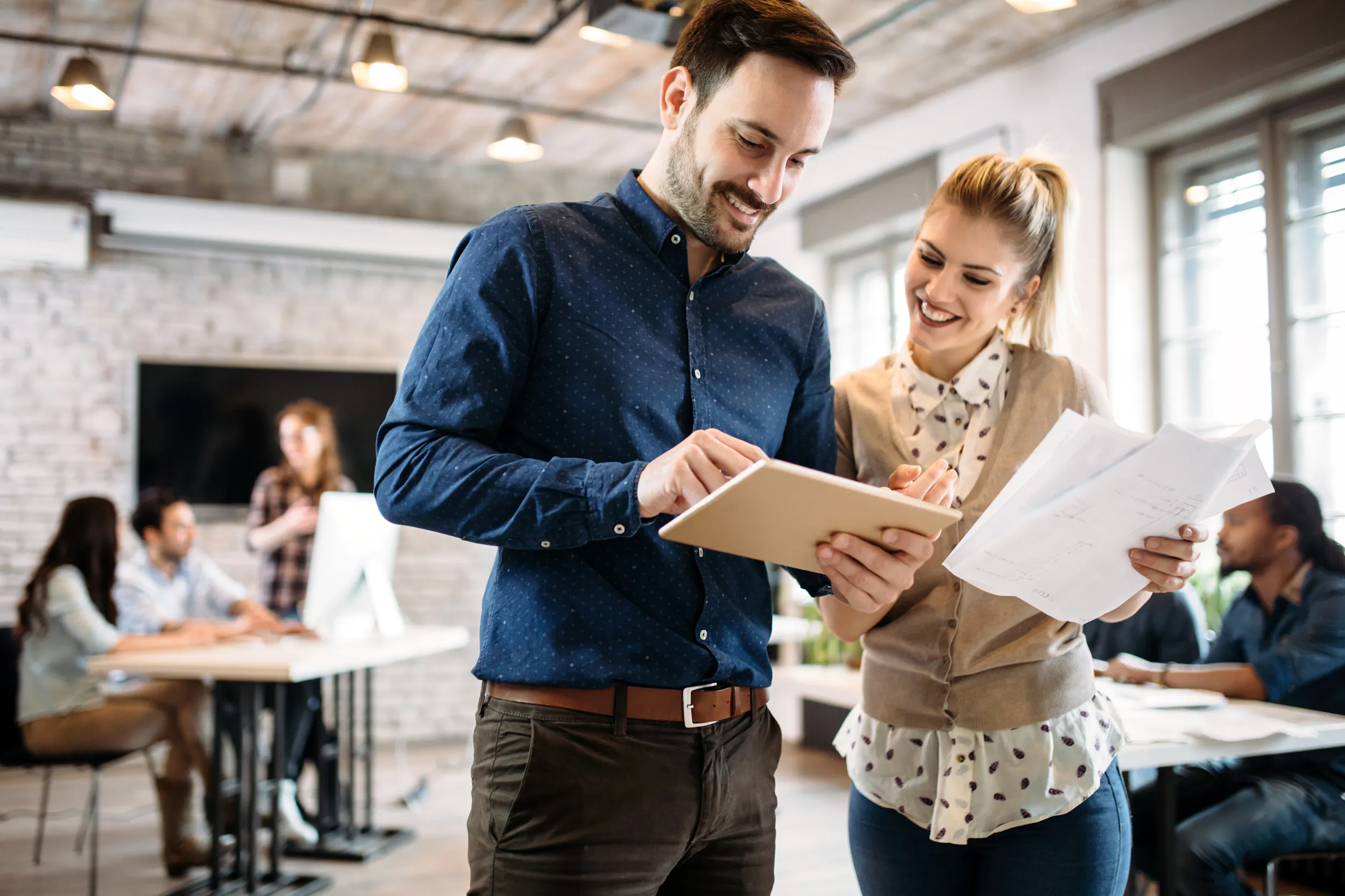 A man and woman stand in an office, one holding a tablet and the other holding papers, engaged in a discussion.